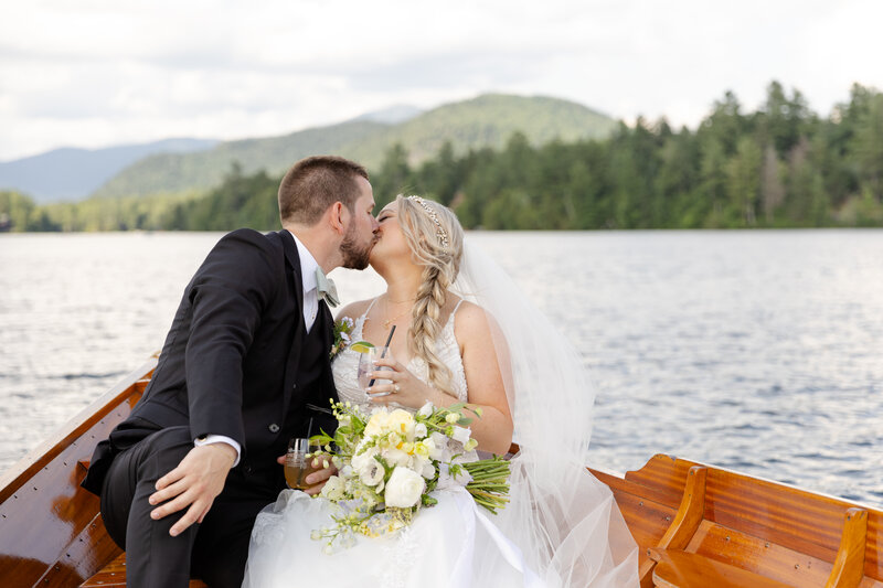 Bride and groom share a kiss on a boat in a lake with the mountains behind them. 