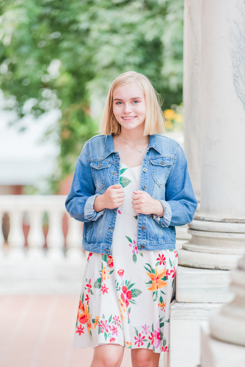 High school senior girl in floral dress and denim jacket smiles on a porch