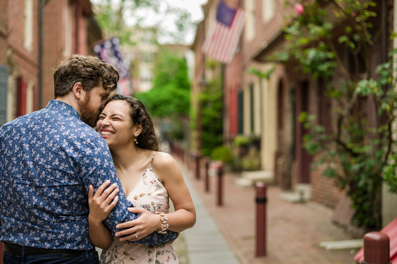 Engaged couple laughing and sharing a joyful moment with the charming, historic background of Elfreth's Alley in Philadelphia.