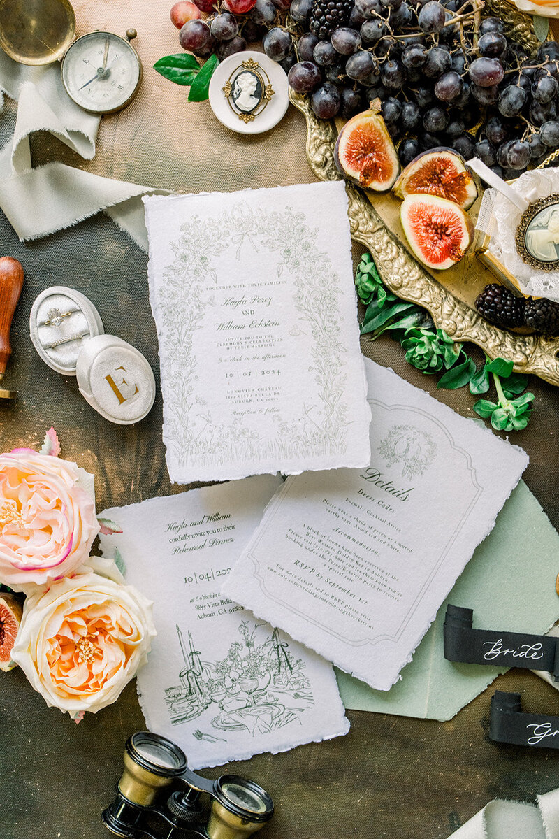 This stunning flatlay showcases a timeless and elegant wedding invitation suite, surrounded by rich details that exude vintage charm. Antique cameos, figs, grapes, and vintage binoculars enhance the opulent setting, complemented by lush peach roses and velvet ribbons. The invitation cards feature delicate letterpress designs on handmade paper, perfectly capturing the romantic atmosphere. This flatlay beautifully sets the tone for a sophisticated wedding, blending old-world elegance with modern refinement, making it an ideal addition to any California wedding inspiration board.