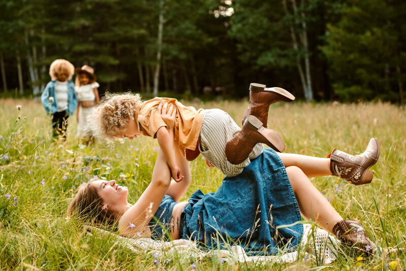 maother holding son in air with other kids playing behind her.