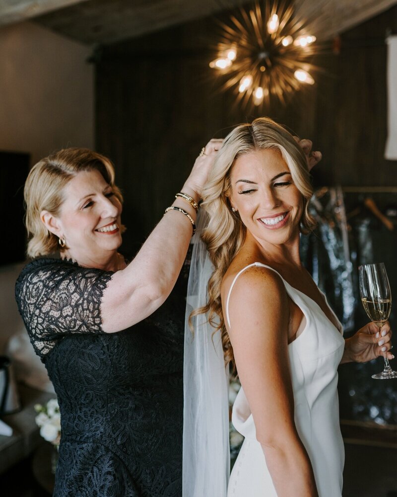 Bride smiling with soft waves and glowing makeup while getting ready, as her mother adjusts her veil—elegant wedding-day glam by Artistry by Jessica.