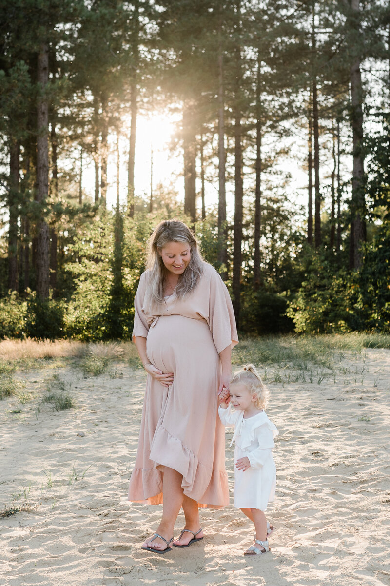 Een intiem moment tussen een zwangere moeder en haar dochter tijdens een fotoshoot in de natuur bij zonsondergang, vastgelegd door Monica Stout Fotografie.