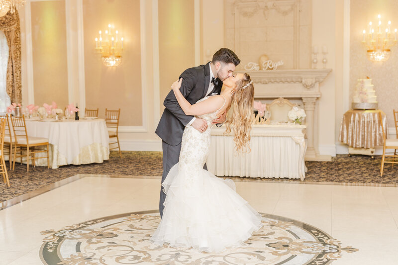A groom dips and kisses his bride while standing in an ornate ballroom