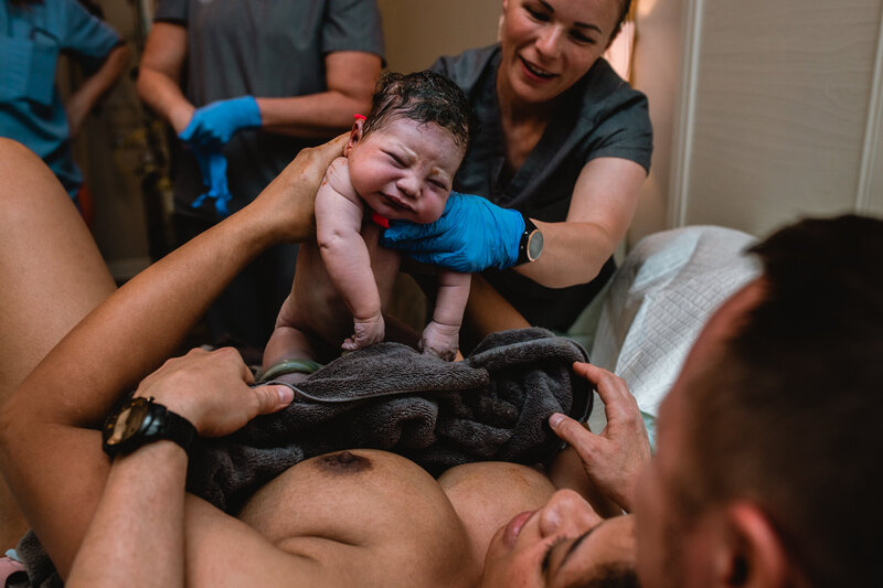 Newborn being lifted to parents’ chest moments after birth while the birth team supports, photographed by a DFW birth photographer