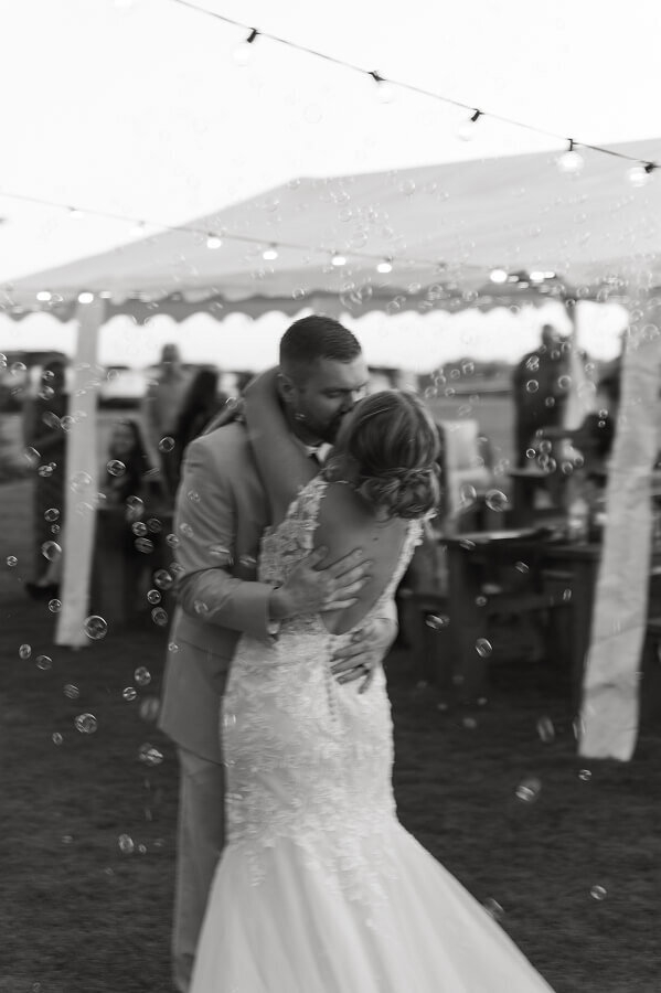 A black and white photo of a bride and groom kissing at their wedding reception. There are bubbles all around them and the photo is slightly blurred. The photo feels like a snapshot in time.