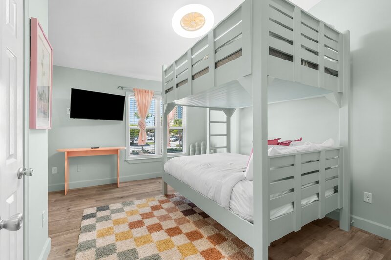Playful kids' bedroom with mint green full-over-full bunk bed, colorful checkered rug, and wall-mounted TV in the Pink Door Surf House.