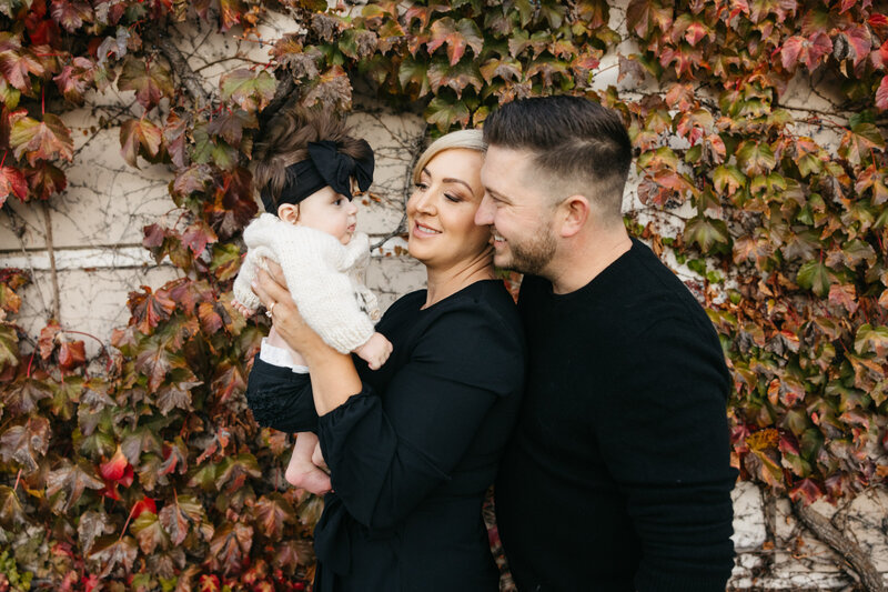 mom and dad holding baby up and smiling while standing in front of ivy wall during the fall