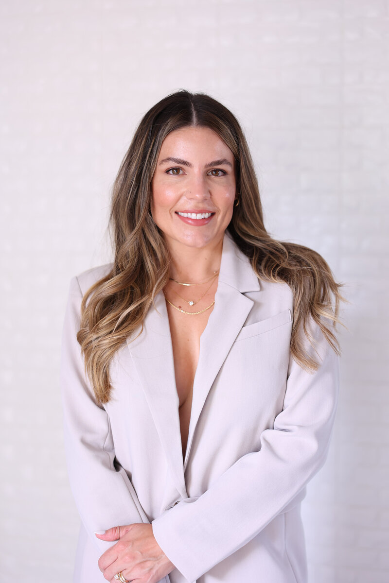 Natalie Bernacchi, a confident woman with long wavy brunette hair, wearing a light blazer and layered gold necklaces, smiling softly in front of a light textured background. 
