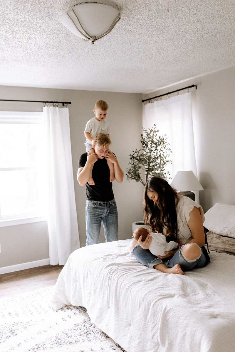 lifestyle newborn session with mama holding baby on bed and dad holding toddler on his shoulders standing next to bed