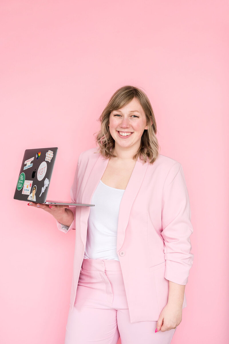 Kat Murphy in a light pink suit holding a laptop standing in front of a pink background