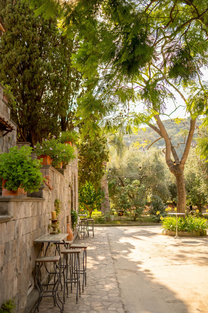 Charming outdoor wedding venue with café tables and chairs on a sunlit stone patio, surrounded by lush greenery.