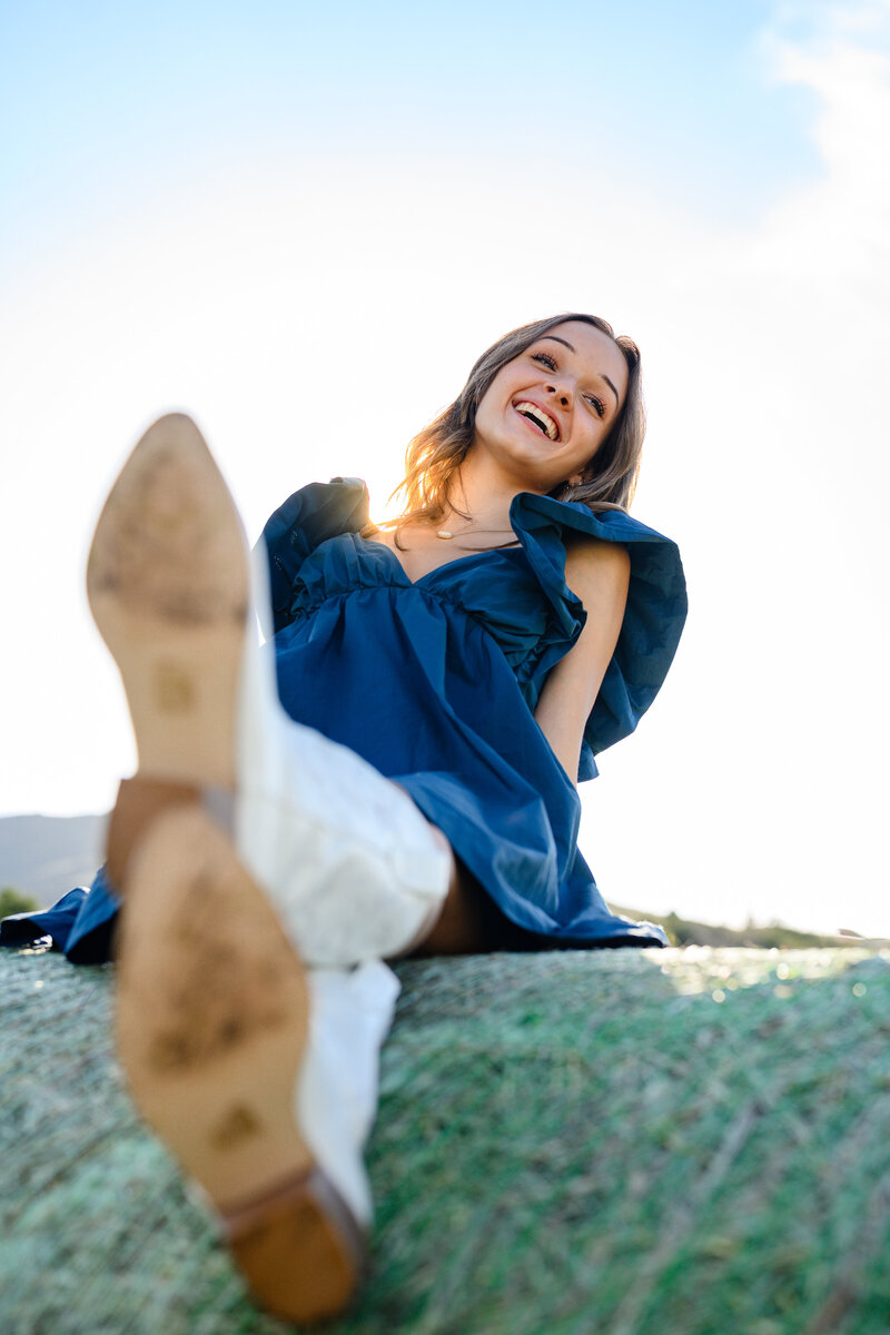 Senior picture photographer near me captures a girl sitting on a hay bale with cowboy boots and the sun flaring behind her