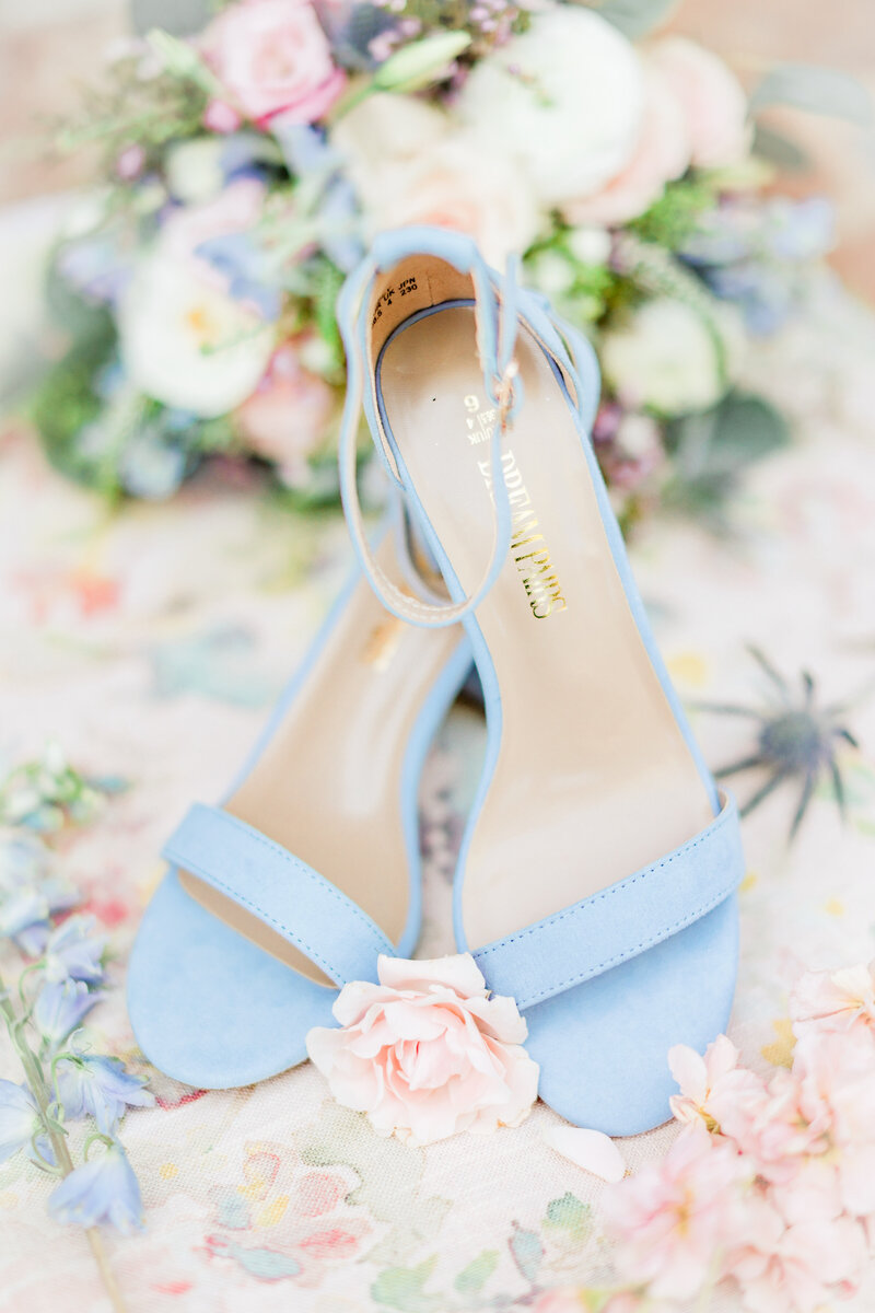 A pair of delicate blue bridal shoes beautifully arranged among soft pastel florals, creating a romantic and timeless detail shot for the wedding day.