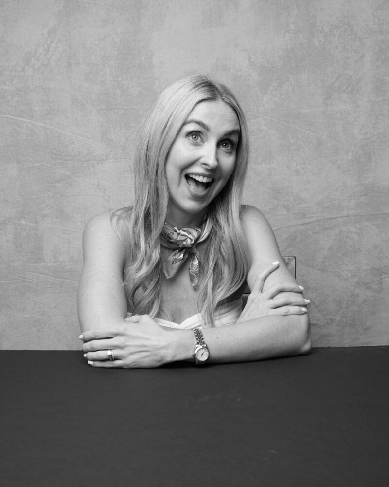 Black and white portrait of a blonde woman sitting at a table, arms crossed playfully as she smiles with wide-eyed excitement. She wears a stylish scarf around her neck, a strapless top, and a watch on her wrist, exuding confidence and charm. A lively, unfiltered moment from 'THIS IS ME – A Photography Experience by Katie Campbell' in Atlanta, GA, celebrating personality and self-expression.