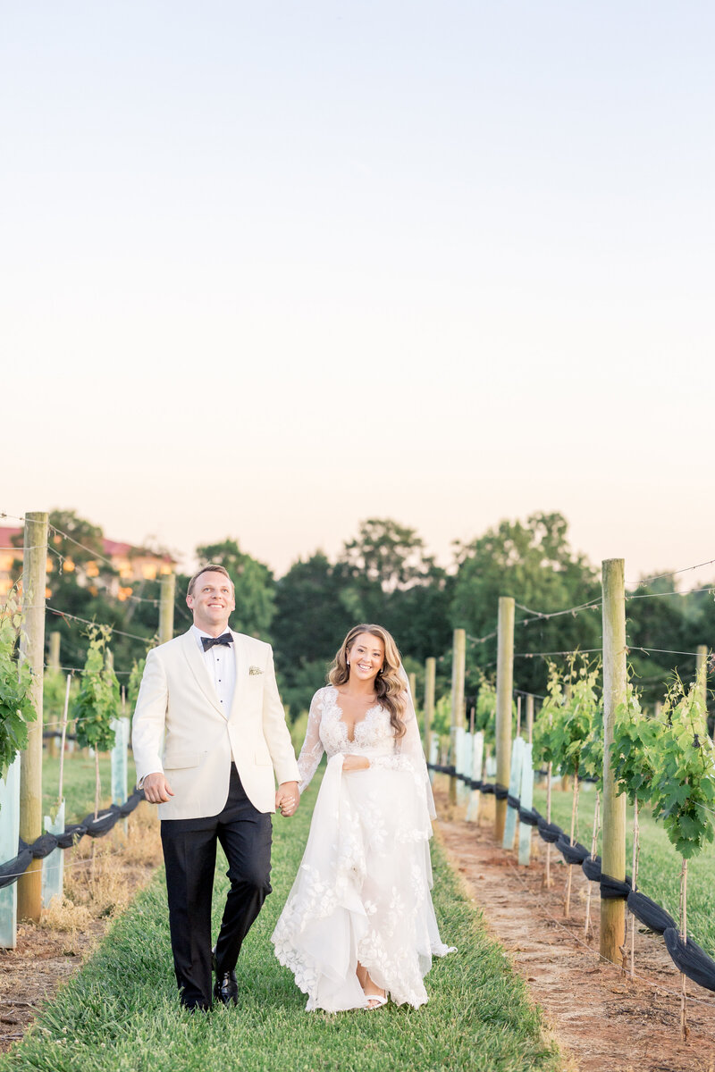 A newlywed couple walks hand in hand through a vineyard, bathed in warm sunset light, exuding romance and timeless charm