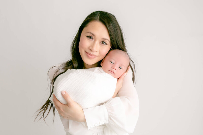 Mom in luxury white dress holding baby wrapped in white. Mom's cheek is resting on baby's face. Mom is looking at the camera with a soft smile on her face and baby's eyes are open and he is looking at the camera.