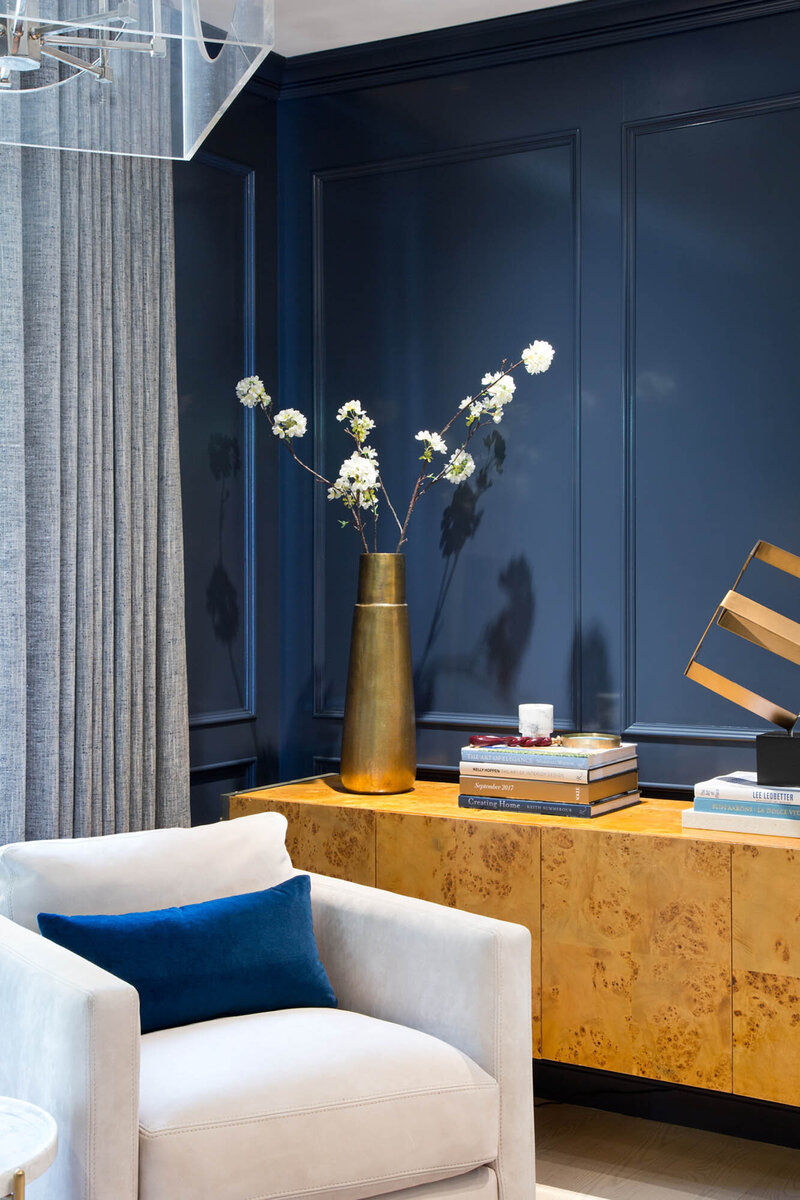 Westbay project, corner of a room with a deep royal blue wall, unique yellow marble patterned console table, a white chair with a blue cushion. There are books and a large gold vase with white flowers on the console table