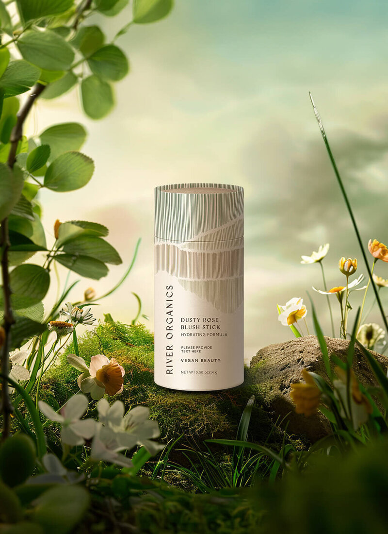 A River Organics product, the Dusty Rose Blush Stick, is displayed in a lush, natural meadow filled with flowers and greenery. The eco-friendly packaging blends beautifully with the organic environment, emphasizing the brand’s commitment to natural beauty.