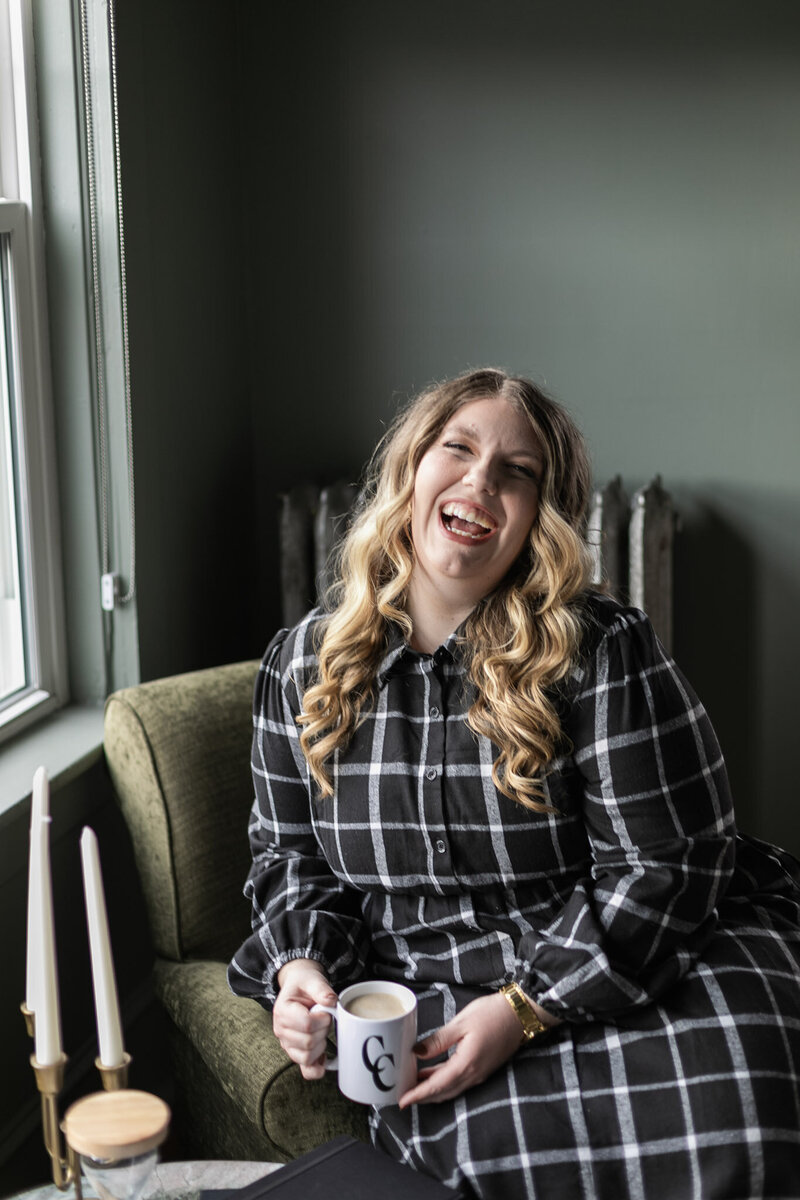 Ashleigh Henry, life coach based in Asheville, North Carolina is pouring an old fashioned in a room at The Blind Tiger, near The Biltmore House and Riverarts Distrct, with green and yellow wall paper behind her, near a window with a small black table holding books and journals. 