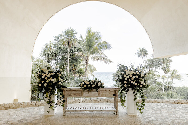 A wedding ceremony setup overlooking the ocean, with lush floral arrangements of white roses and greenery on either side of a wooden altar, framed by an arched entrance with palm trees in the background. Keywords: destination wedding planning, luxury destination weddings, wedding ceremony decor.