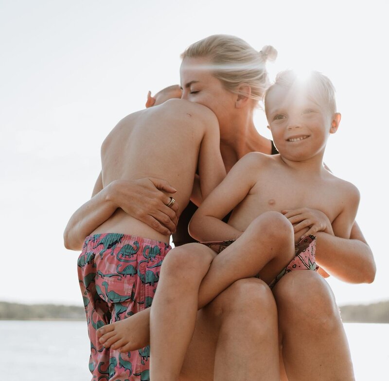 Lifestyle family photo of a mother embracing her two young sons at the lake during golden hour, capturing a candid moment of love and connection.