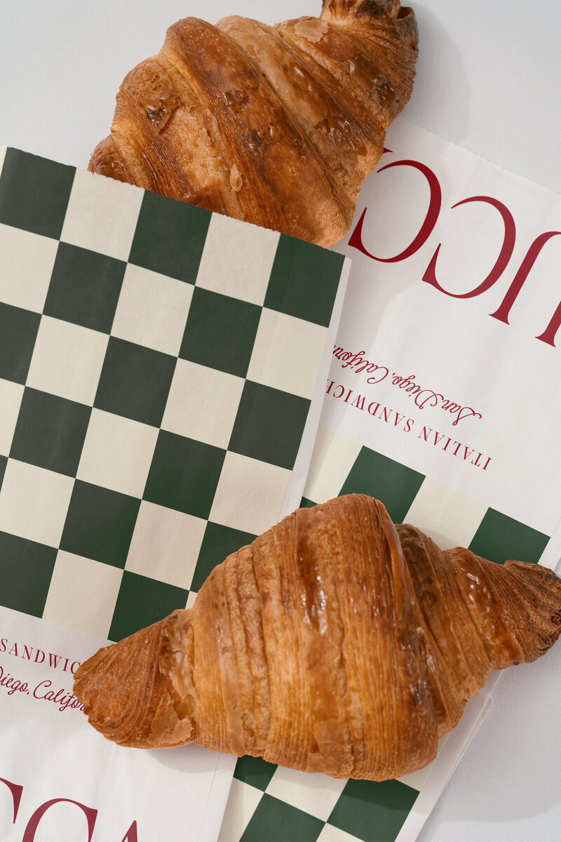 LUCCA Italian Sandwich Shop branded paper bags with croissants, featuring green checkerboard pattern and elegant packaging design.