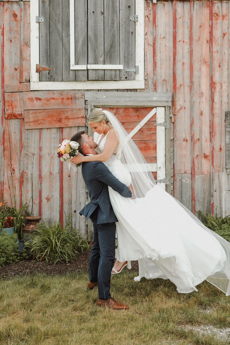 Groom lifts his bride on their wedding day in front of rustic barn wedding venue