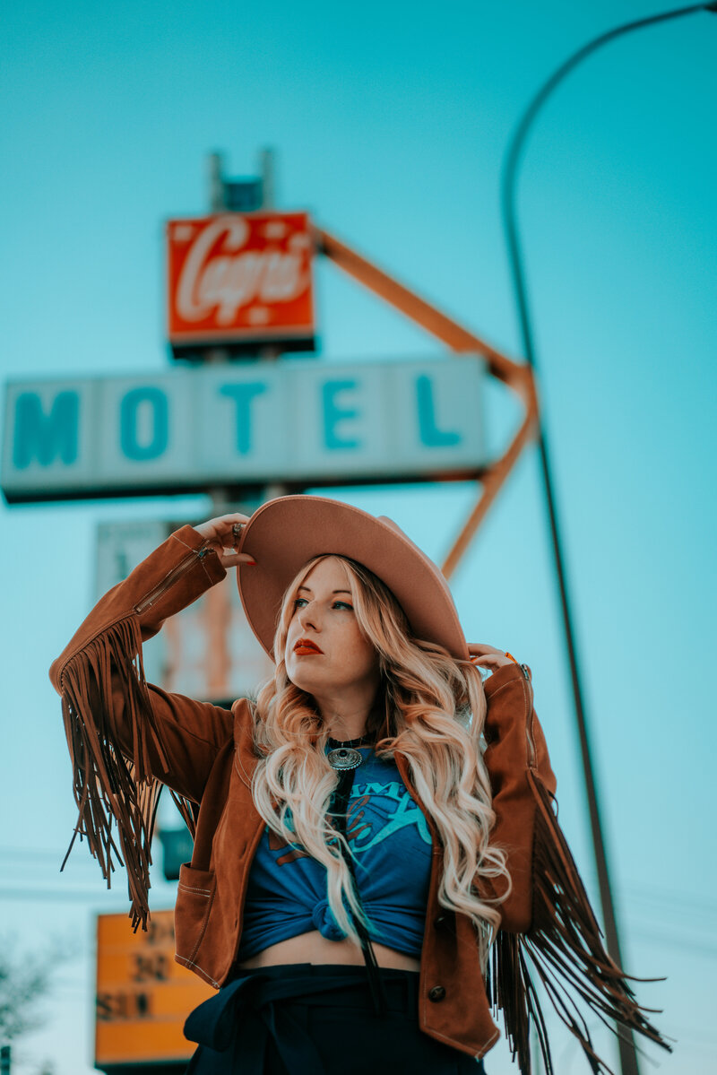 Photo of blonde woman in front of motel wearing fringe jacket and cowboy hat