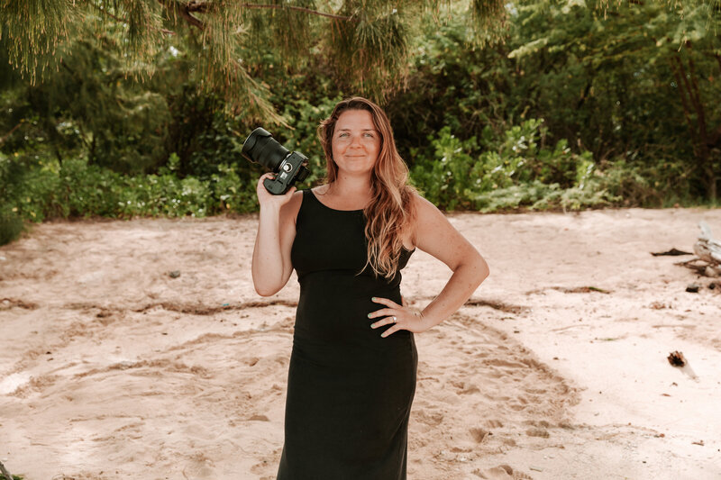 Sierra owner of sierra bertini photography. I am wearing a long black dress on the beach with my hand on one hip and holding my camera with my other hand. I am standing on the beach with a green background and sand