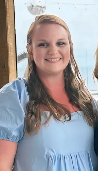 A portrait of a destination wedding planner with long wavy hair, wearing a light blue blouse and smiling, taken indoors with a window view of the outdoors in the background.