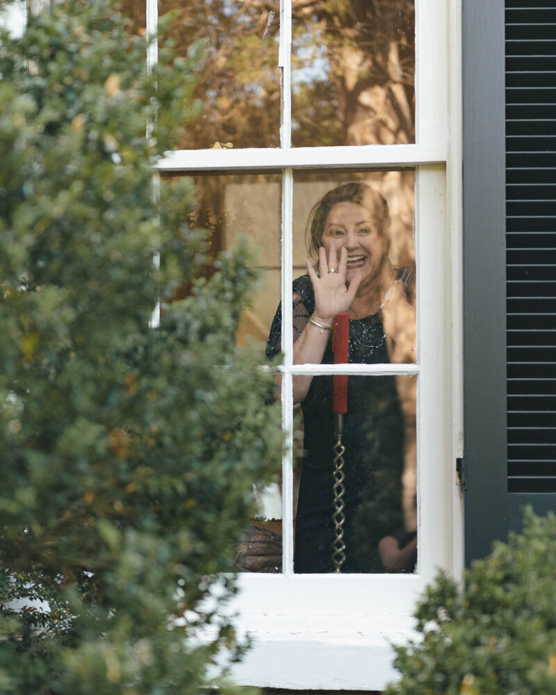 A mother of the groom looks out of the window as she watches a first look. She looks at the camera and waves, showing the documentary-focus of this photographer.