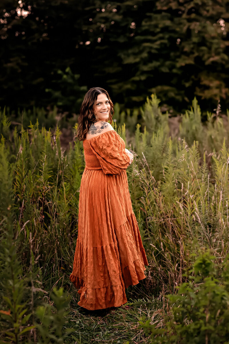 Woman wearing a long orange dress is standing in a field of tall wild grass. She is hugging onto herself and smiling.