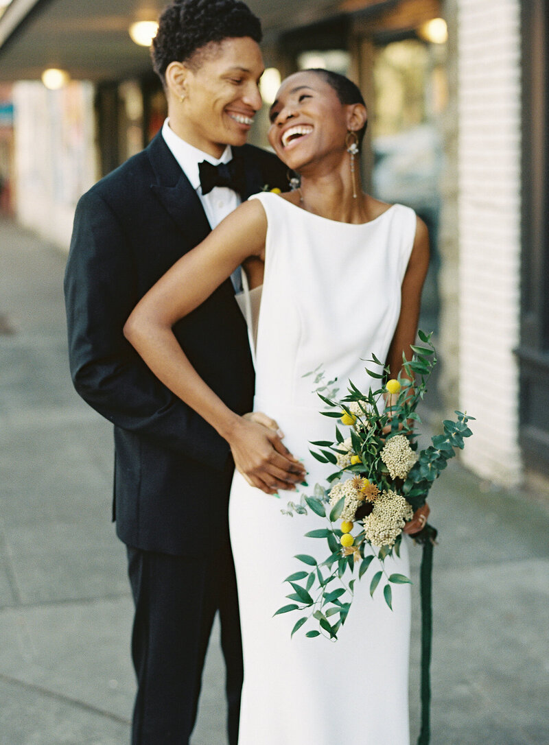 Photo of a groom in a black tuxedo embracing his bride from behind. The bride is wearing a modern and simple style wedding dress and has a close-crop hair cut.