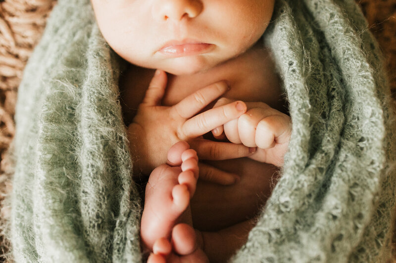 Family of 4.  Parents are standing back to back .  Mom is holding her 2 year old and looking at her and smiling.  The 2 year old is loking directly ta the camera with an open mouthed smile.  Dad is holding newborn baby girl. He is also looking directly at the camera and smiling.