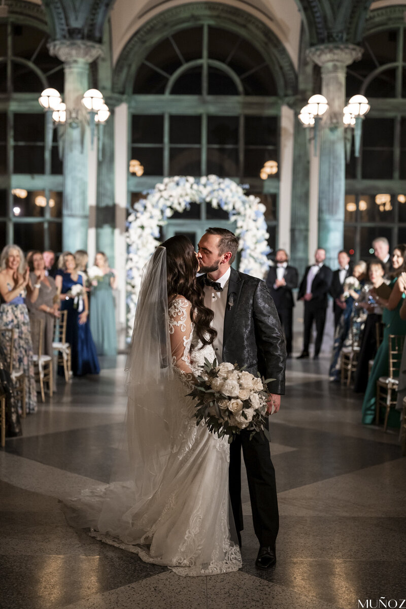 A bride and groom share a kiss in front of a floral wedding arch, surrounded by family and friends, embodying the romance of a luxury wedding ceremony.