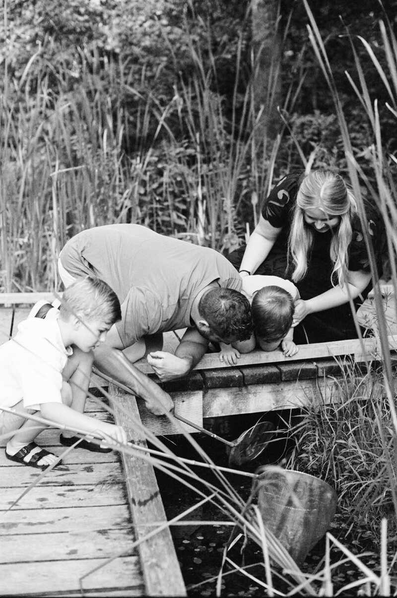 A family leans over a wooden bridge, observing the water below. A man holds a net, while a woman and two children watch intently, capturing family film moments in Enniskillen by family film photographer Jennifer van Son.