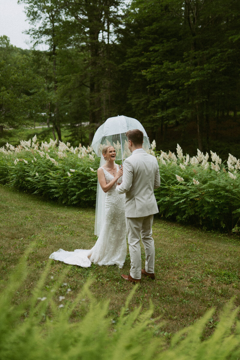 Bride in a wedding dress holding an umbrella and groom in a cream-colored suit smiling at each other during their first look at Lakefalls Lodge in Stoddard, New Hampshire.