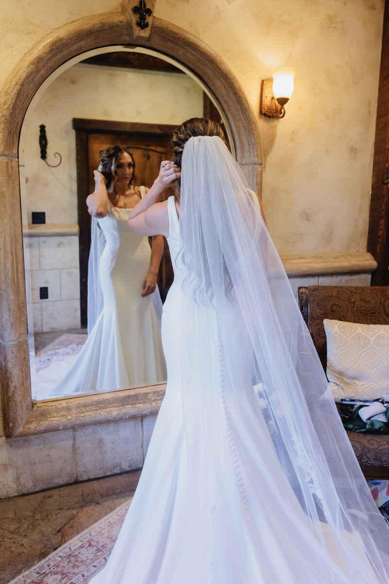 A bride stands in front of a large mirror adjusting her veil