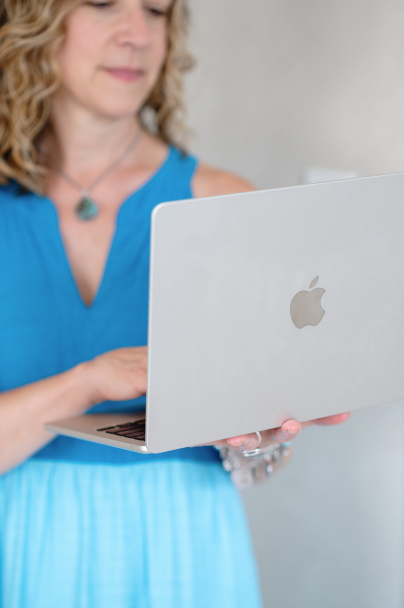 A woman in a blue dress, with curly hair, uses a silver laptop with an Apple logo. Her expression is focused and the tone is professional.