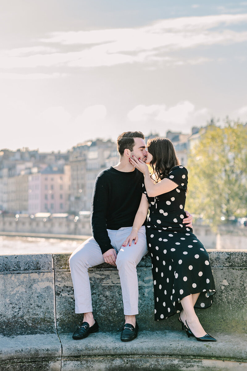 Man and woman sitting on a bridge near Pont Neuf on Île de la Cité during a Paris proposal — the woman in a polka dot dress gently holding the man’s face.