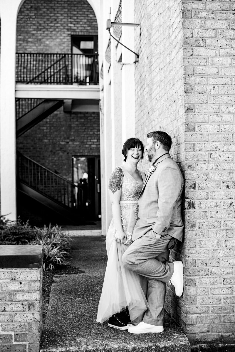 Black and white photo of groom leaning against wall and bride laughing with him