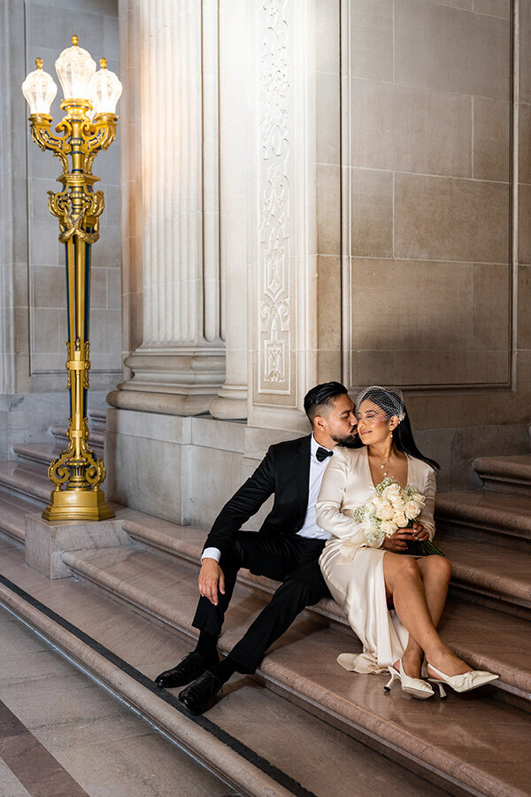Professional San Francisco Bay Area wedding photography featuring intimate portrait as bride in elegant champagne dress with birdcage veil and groom in black tuxedo share tender moment on San Francisco City Hall's marble steps with neoclassical columns, ornate golden lamp post, and classical architecture creating timeless elegance