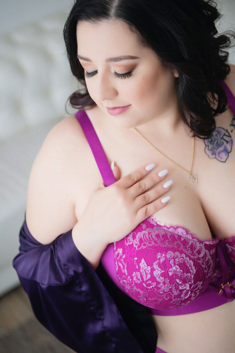 Woman in a purple lace bra and robe, resting her hand on her chest, looking down with closed eyes. She has a floral tattoo and wears a necklace.