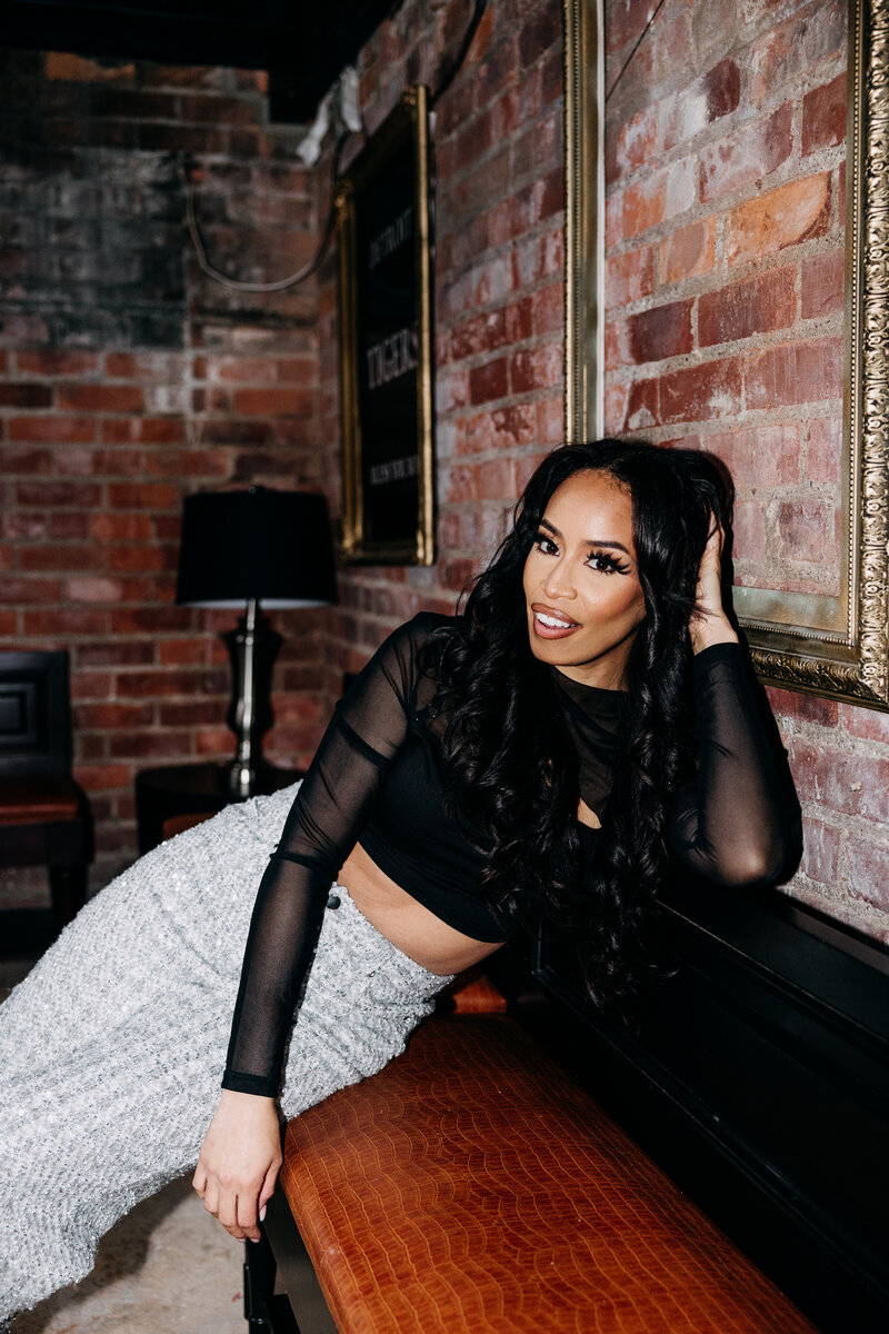Flash photo of Tiara in a black top and sparkly pants sitting on a bench leaning against a brick wall