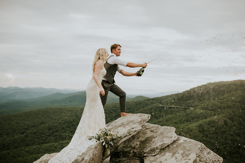 bride and groom standing on cliff opening a champagne bottle with mountains in background