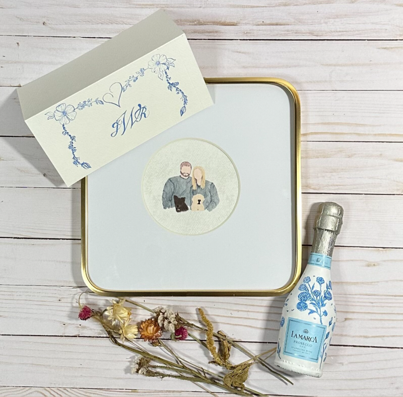 A flatlay picture on top of white barnboard, of a hand-made wedding card with a three initial monogram, a miniature bottle of prosecco that is painted white all over, with blue flowers painted on it. A bouquet of dried flowers to go in the bottle when the prosecco is gone. The hero gift is a mini faceless watercolor of the couple and their two dogs.