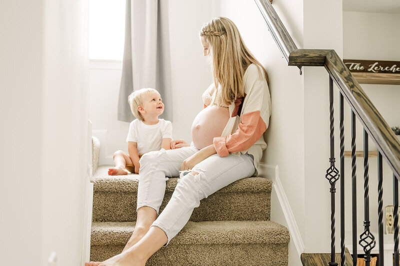 pregnant mother sitting on the landing of her stairs with her belly showing next to her two year old son at home by york pa photographer