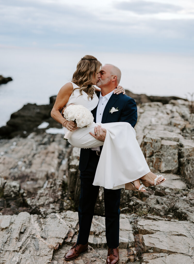 Groom stands on rocks in ocean and holds bride and kisses her at The Cliffhouse in Maine