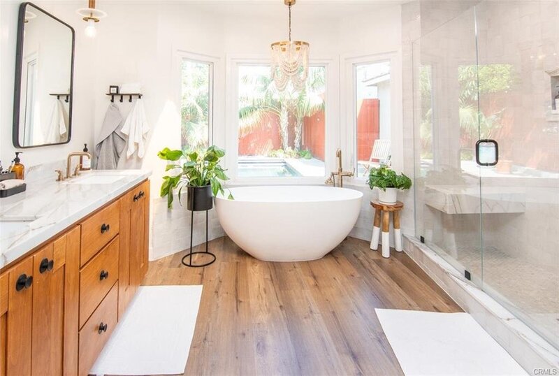 Bright master bathroom with soaking tub, bay window, chandelier, hardwood floors, white walls, potted ferns, and a full length glass shower.